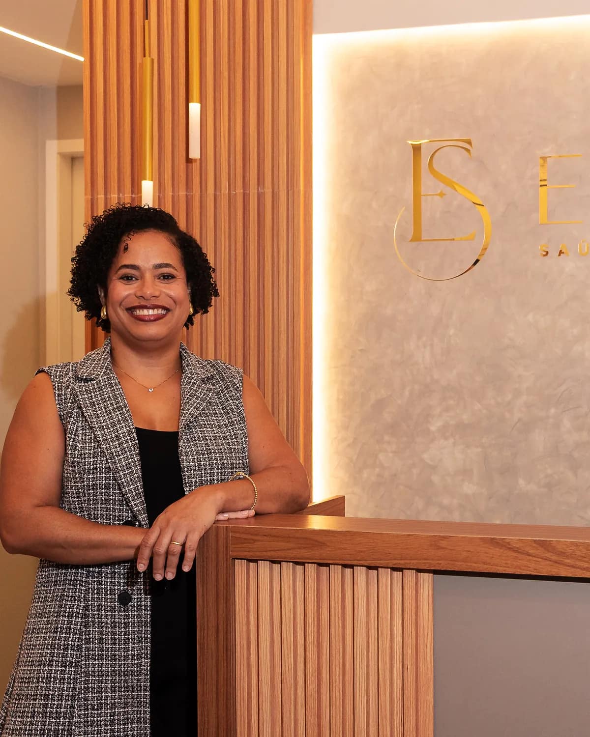 Viviane Maia recebendo no balcão da recepção da Essenza, com o monograma dourado ao fundo